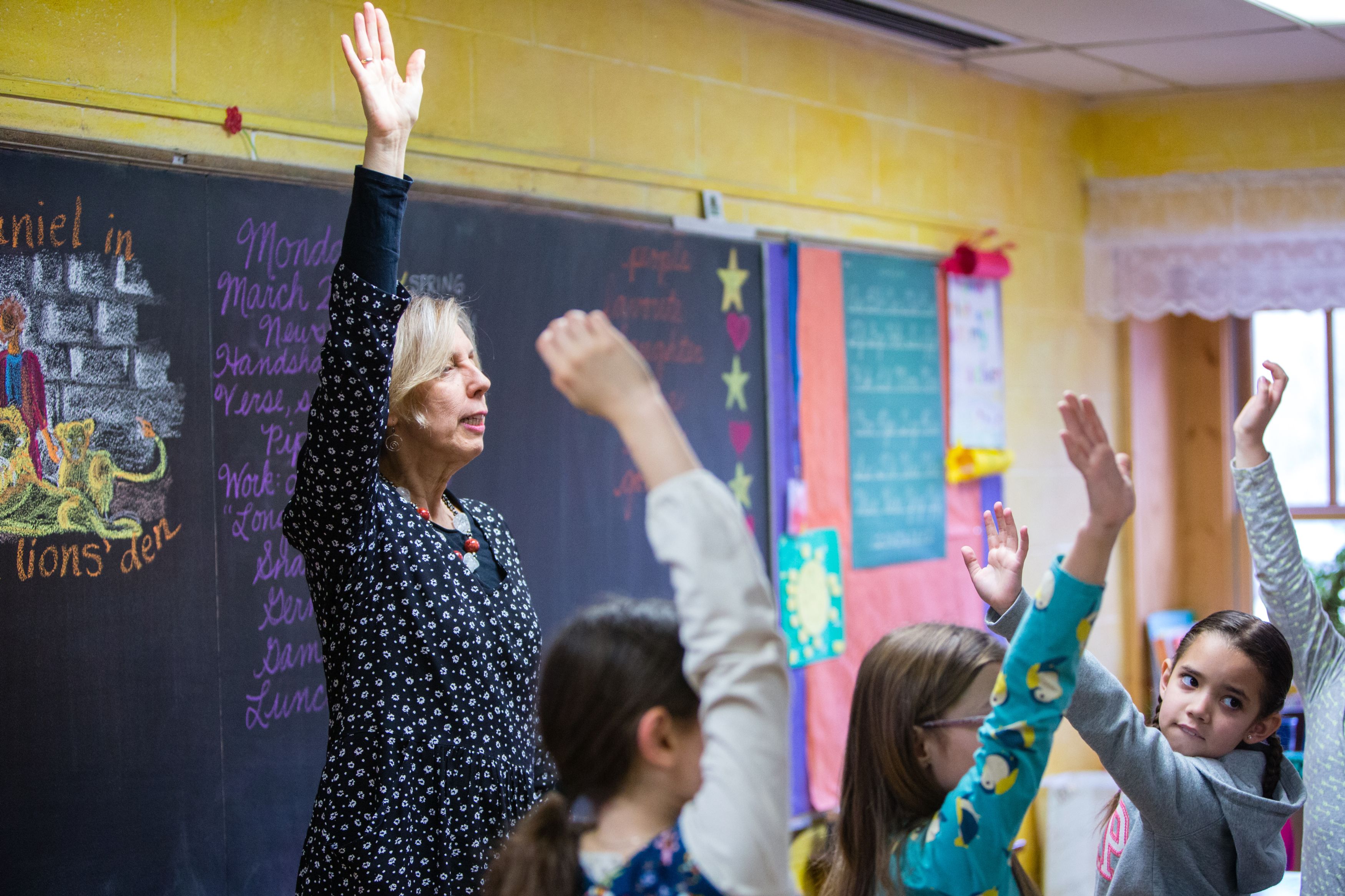 Teacher and students raising hands at front of classroom